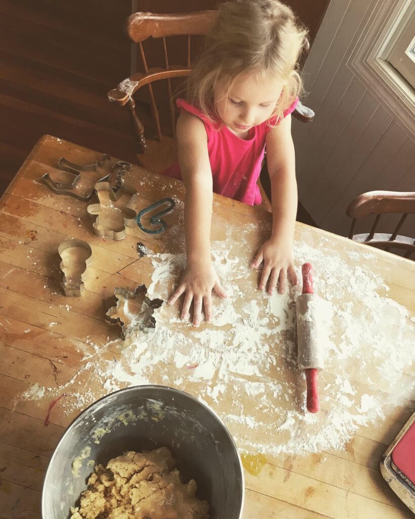 little girl making sugar cookies at kitchen table with dough, flour and rolling pin