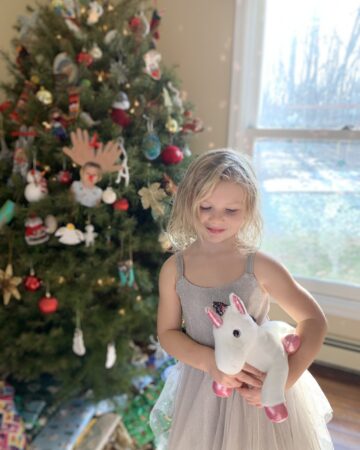 little joy-filled girl in front of simple Christmas tree in holiday dress with unicorn stuffed animal